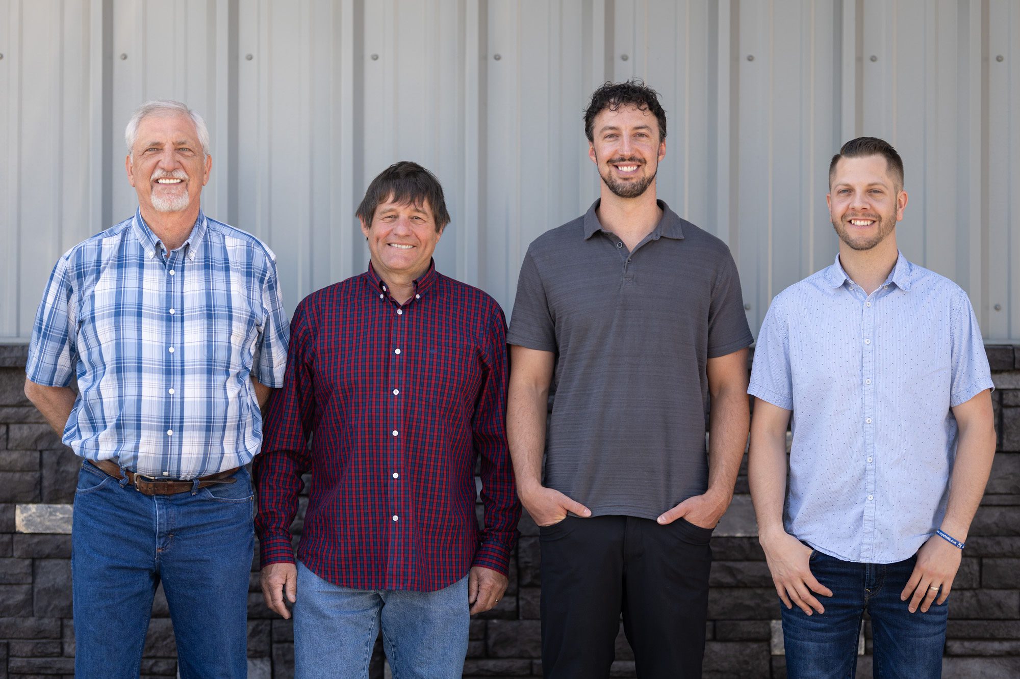 Group Photo Four men stand in a row, smiling in front of a corrugated metal and brick wall. Each wears casual attire: two in button-up shirts, one in a polo, and one in a plaid shirt.