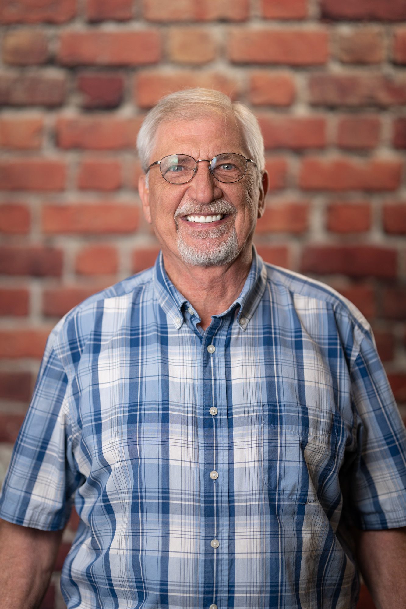 Mike Hohl Elderly man with glasses and a short white beard smiling, wearing a blue plaid shirt, and standing in front of a brick wall.