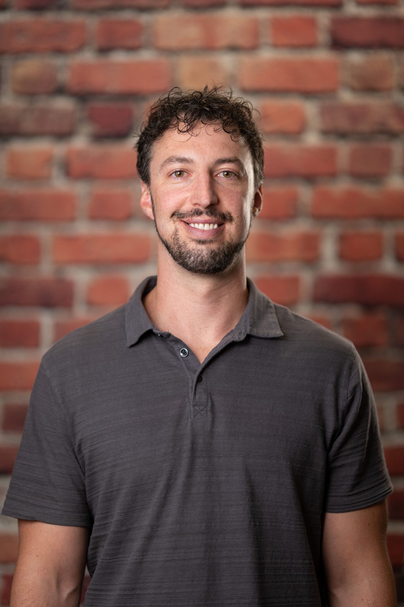 Tyler Cavanaugh A man with short curly hair and a beard is smiling and wearing a gray polo shirt, standing in front of a brick wall.